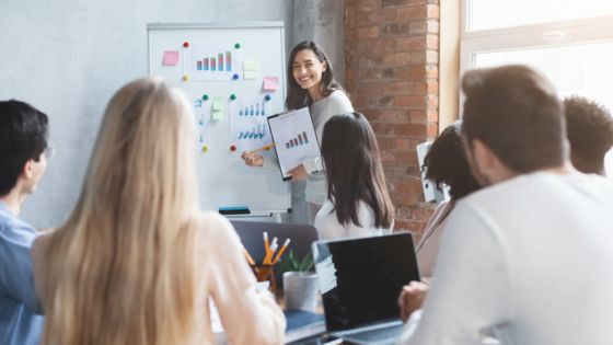 Team meeting with a woman presenting charts on a whiteboard in a modern office setting.