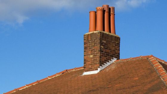 Brick chimney with multiple clay flue pipes on a red-tiled roof against a clear blue sky.