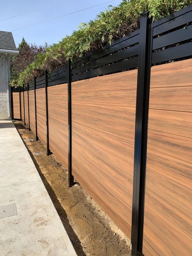 Modern wood and metal privacy fence with greenery on top, alongside a concrete walkway.