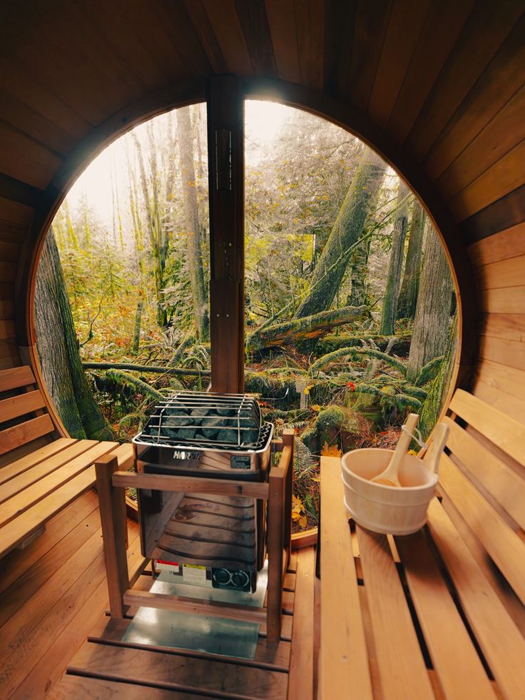 Wooden sauna with forest view through round window, featuring seating and a bucket with ladles.