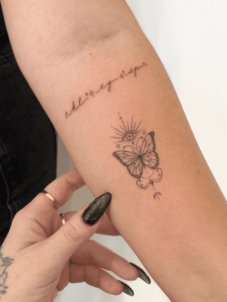 Delicate butterfly tattoo on forearm with an eye above, surrounded by line art and script letters on skin.