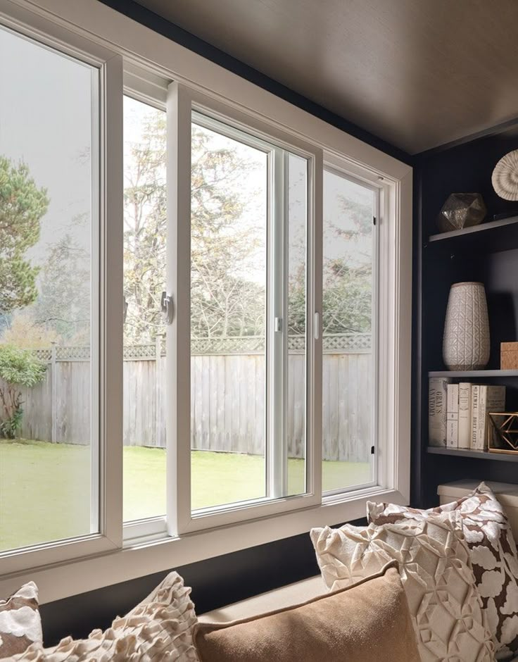 Modern living room with large windows overlooking a backyard, featuring cozy seating and decorative shelves.