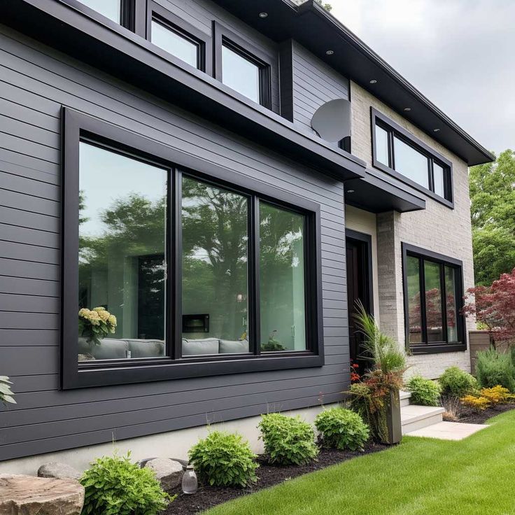 Modern house exterior with large black-framed windows, gray siding, and landscaped green garden.