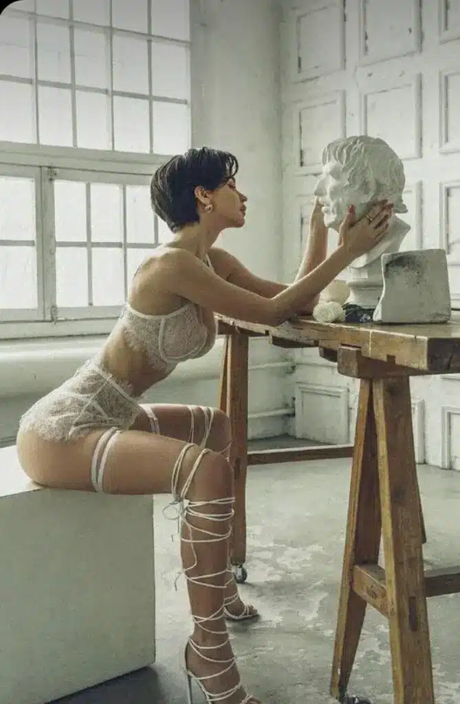 Woman in lace outfit admires marble bust on a wooden table in a bright studio setting.