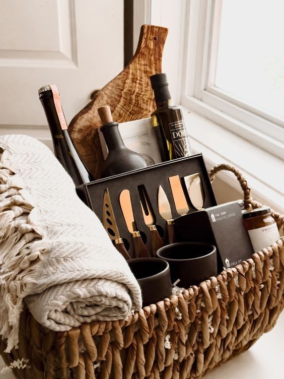 Gift basket with wine, cheese knives, olive oil, and a woven blanket by the window, ideal for cozy indulgence.