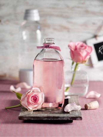 Glass bottle of pink rose water on wooden tray with pink roses and petals, against a soft blurred background.