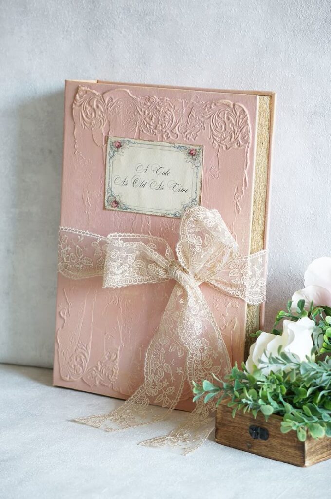 Elegant pink book with lace ribbon, embossed floral design, and A Tale As Old As Time text.