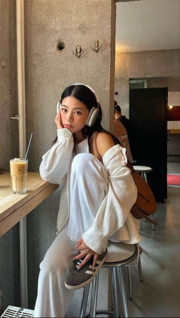 A person in a cozy cafe setting, wearing headphones and stylish white outfit, sipping iced coffee.