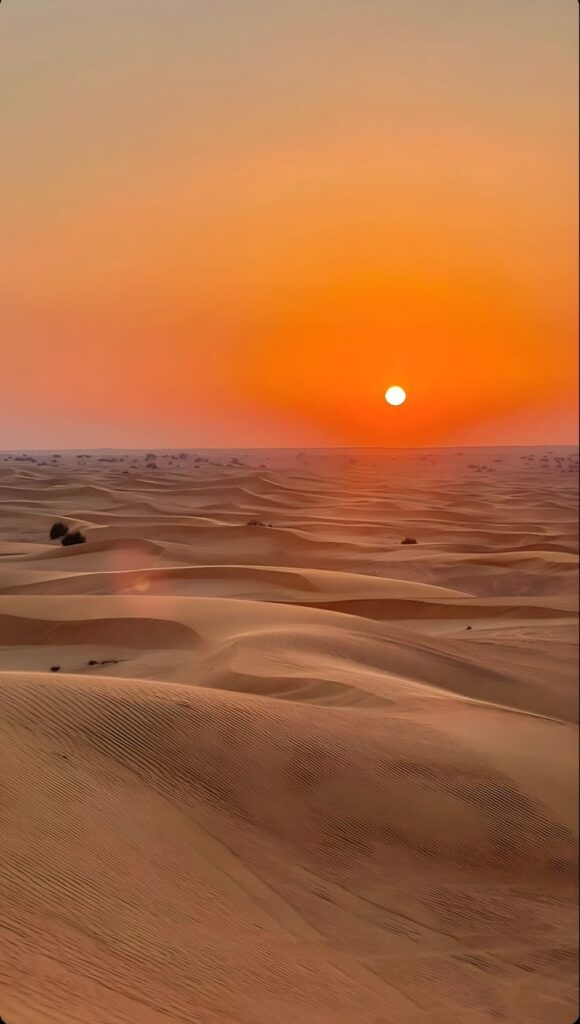 Sunset over expansive desert dunes with orange sky and serene landscape.