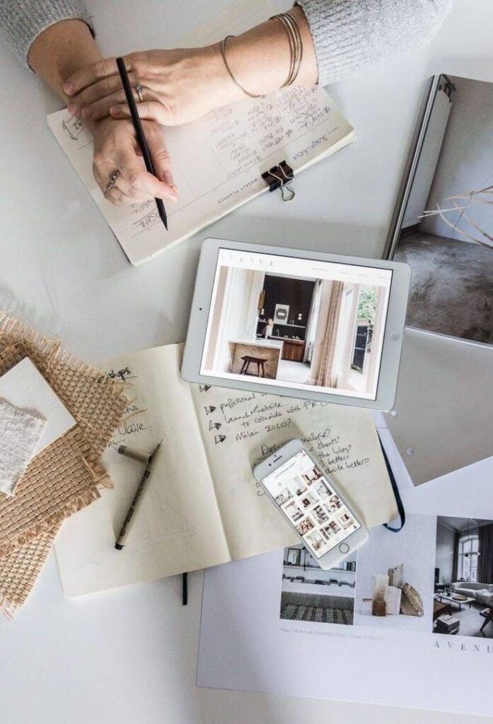 Person planning interior design with tablet and smartphone on a desk, surrounded by notes and fabrics.