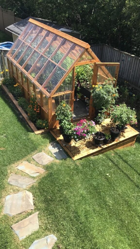 Sunny backyard greenhouse with flowers and plants, wooden structure, clear panels, and stone path on green grass.