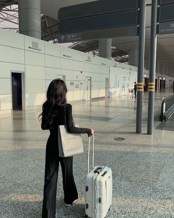 Woman in airport with suitcase, wearing black outfit, walking towards terminal, traveling light with bag and luggage.