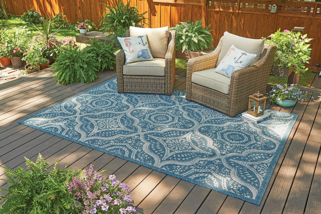 Outdoor patio with wicker chairs, blue patterned rug, plants, and cushions. Cozy backyard seating area.