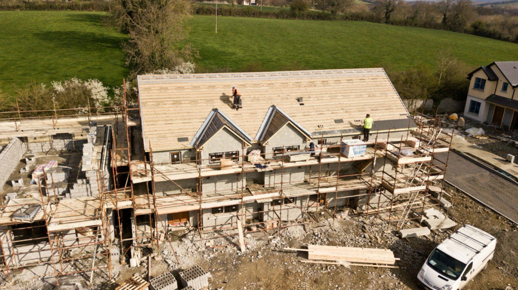 Aerial view of house under construction with scaffolding and workers, set against green fields and countryside.