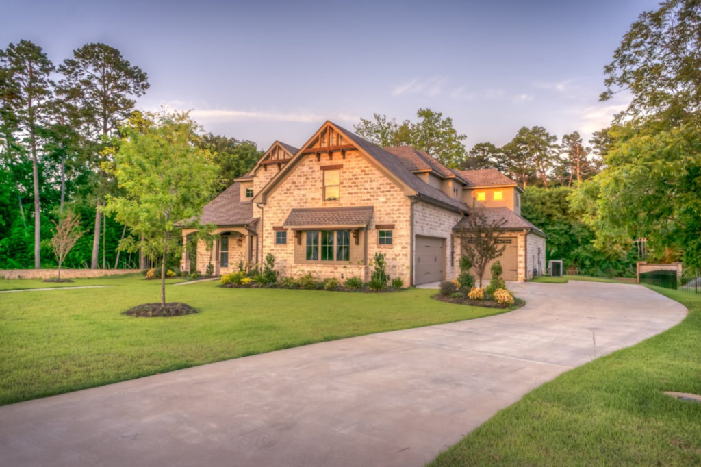 Charming stone house with lush green lawn and curved driveway, surrounded by trees under a clear sky.