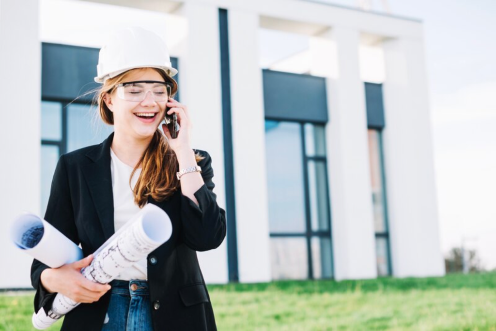 Smiling female architect in hard hat on phone, holding blueprints at construction site.