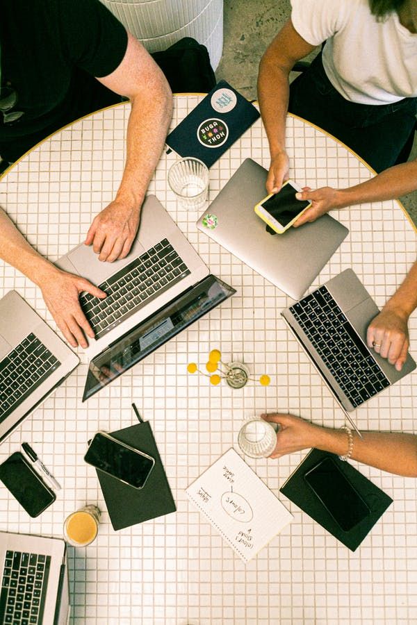 Team collaborating with laptops and smartphones around a table, sharing ideas and drinks in a casual workspace setting.