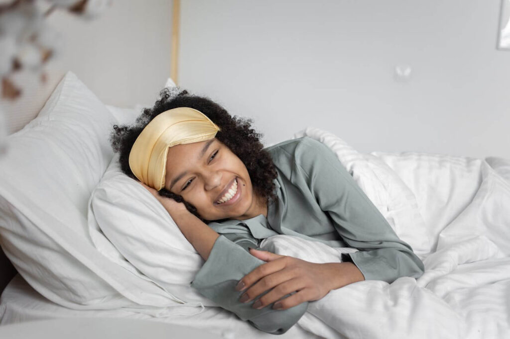 Smiling woman wearing a sleep mask, lying in bed, enjoying a restful morning in cozy sheets.