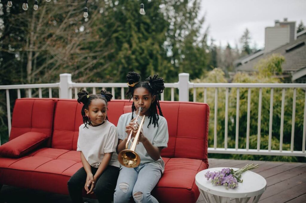 Two children on a red outdoor sofa, one playing a trumpet, with a white table and flowers nearby.