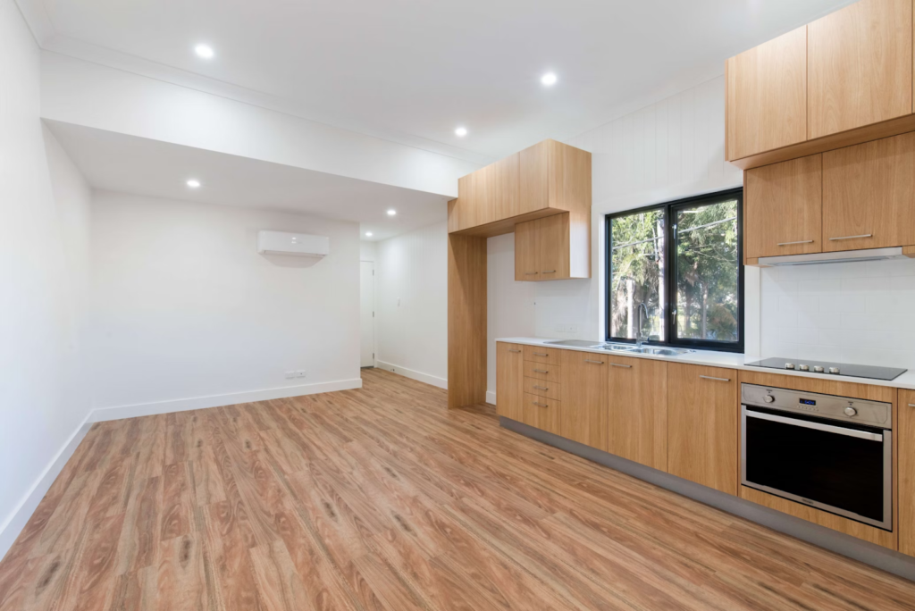 Modern kitchen with wooden cabinets and flooring, featuring a built-in oven and large window for natural light.