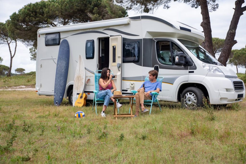 Couple relaxing by camper van with surfboards, guitar, and chairs in a scenic outdoor setting.