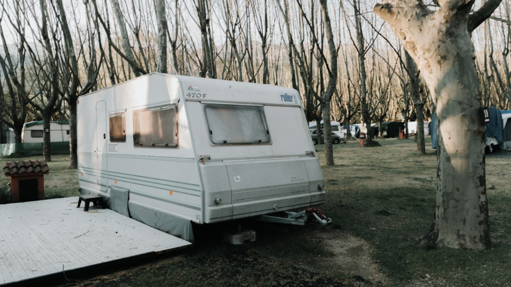 White caravan parked in a wooded campsite, surrounded by trees and sunlight filtering through branches.