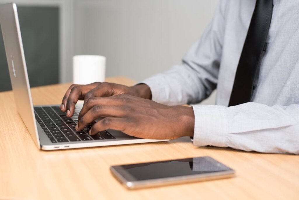 Person typing on a laptop at a desk with a smartphone, wearing formal attire.