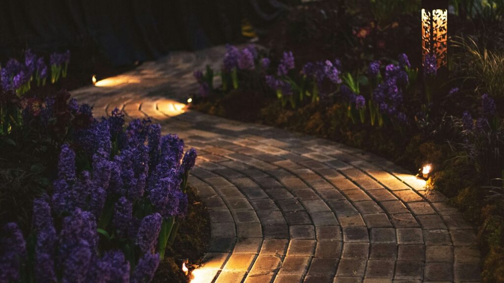 Illuminated brick path surrounded by vibrant purple flowers at night.