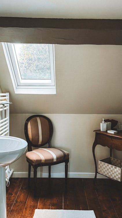 Cozy bathroom attic space with a window, vintage chair, and wooden table, featuring natural light and neutral tones.