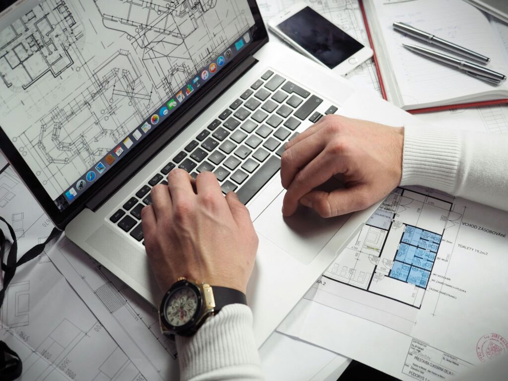 Architect working on floor plans using a laptop, surrounded by blueprints and stationery.