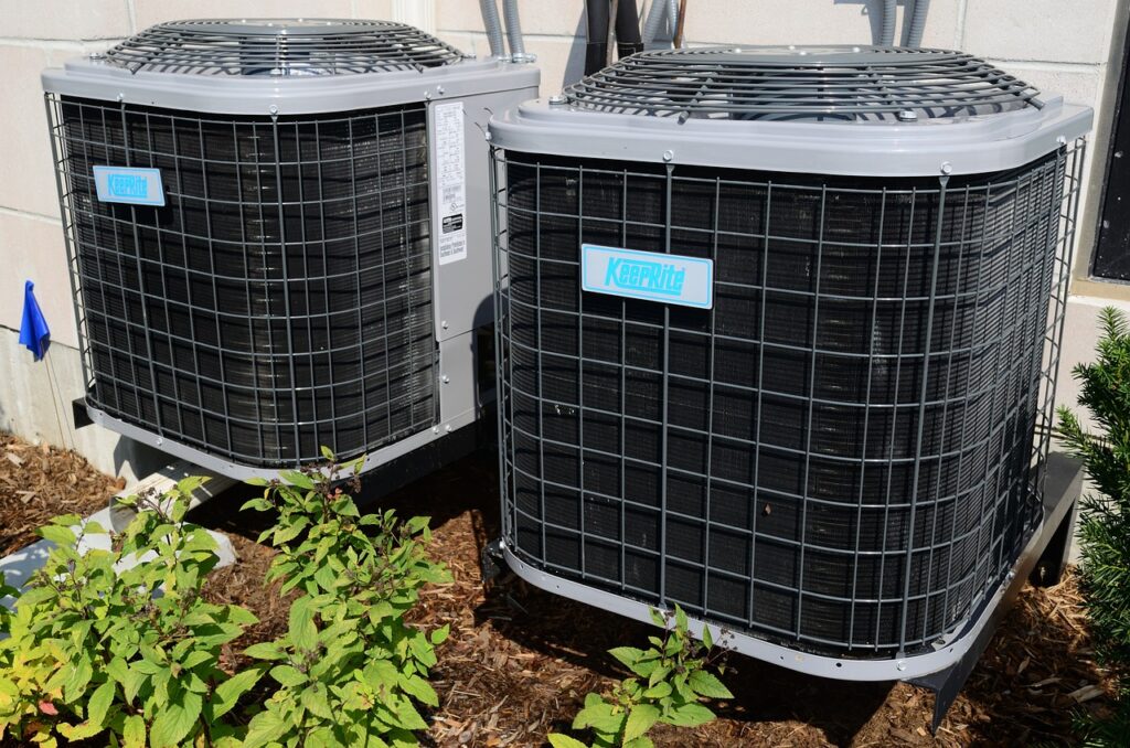 Dual outdoor air conditioning units with protective grilles beside a building, surrounded by plants.