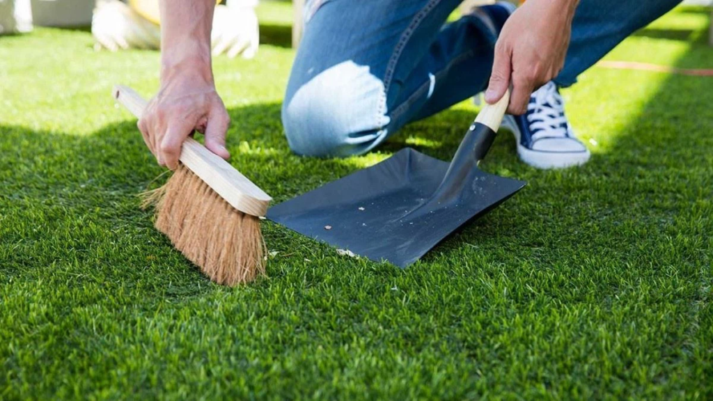 Person cleaning artificial grass with broom and dustpan in a garden setting, showing lawn maintenance.