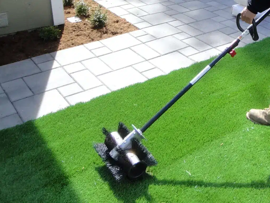 Artificial turf installation with a power broom on a sunny day in a backyard setting.