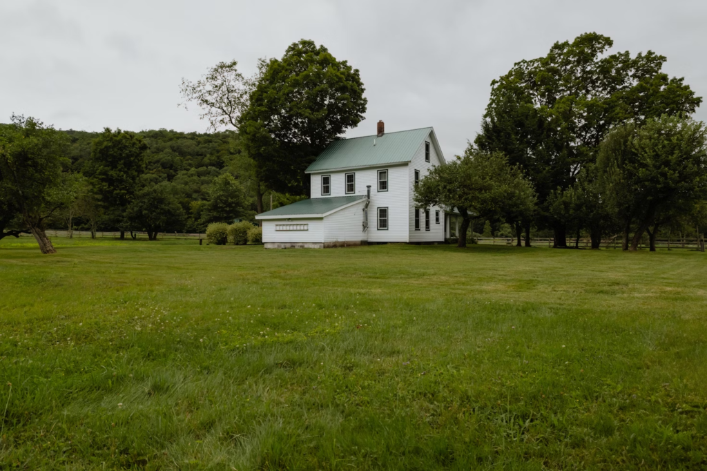 White farmhouse with green roof surrounded by lush trees and open grass field under cloudy skies.