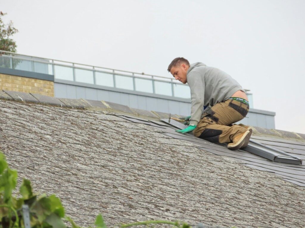 Roofer in protective gear repairs roof tiles on a residential building on a cloudy day.