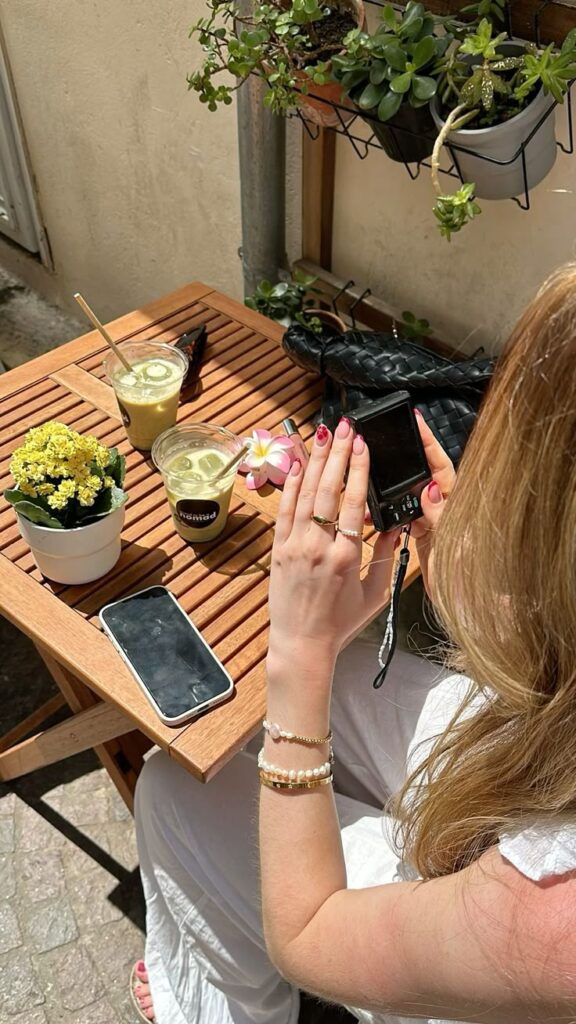 Person photographing drinks and flowers on an outdoor cafe table with a phone and plants nearby.