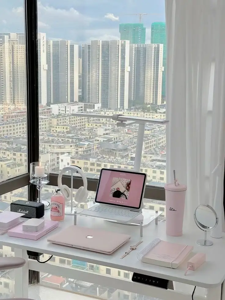 Stylish home office setup with pink accessories, laptop, headphones, and city view through large windows.