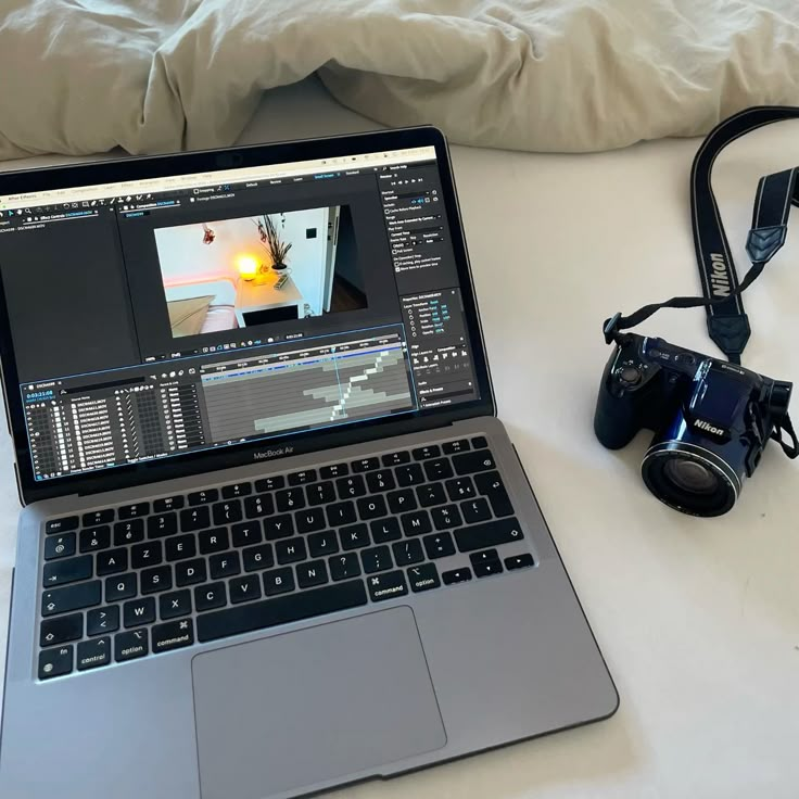 A MacBook Air showing video editing software next to a Nikon camera on a bed.