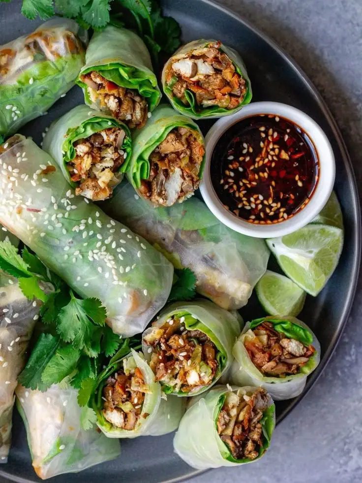 𝐂𝐇𝐈𝐂𝐊𝐄𝐍 𝐒𝐏𝐑𝐈𝐍𝐆 𝐑𝐎𝐋𝐋𝐒🌱 Fresh spring rolls with chicken, veggies, cilantro, and dipping sauce on a dark plate, garnished with lime wedges. | Sky Rye Design Fresh spring rolls with chicken, veggies, cilantro, and dipping sauce on a dark plate, garnished with lime wedges.