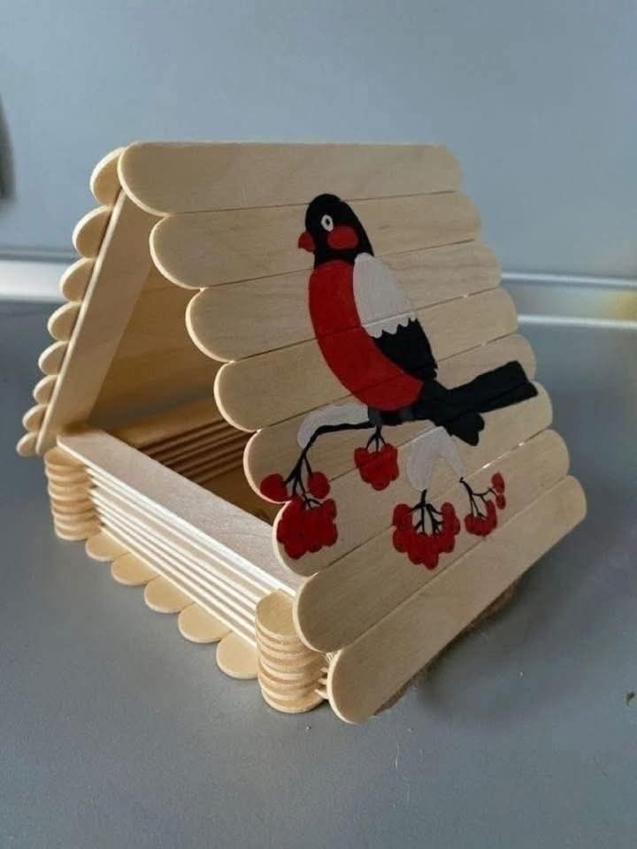 Popsicle stick birdhouse with painted red bird and berries design on the side, resting on a smooth surface.