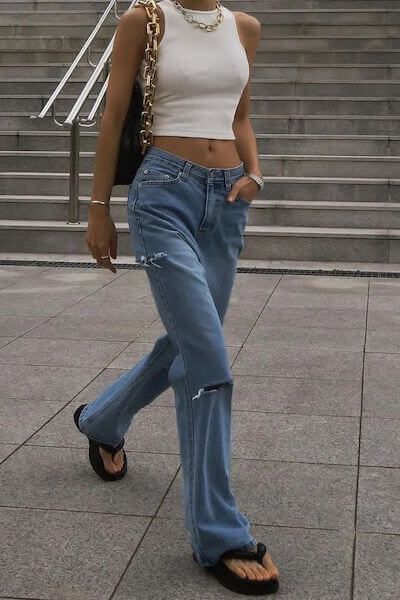 Fashionable woman in ripped jeans and white crop top, accessorized with a handbag and necklace, walking confidently outdoors.