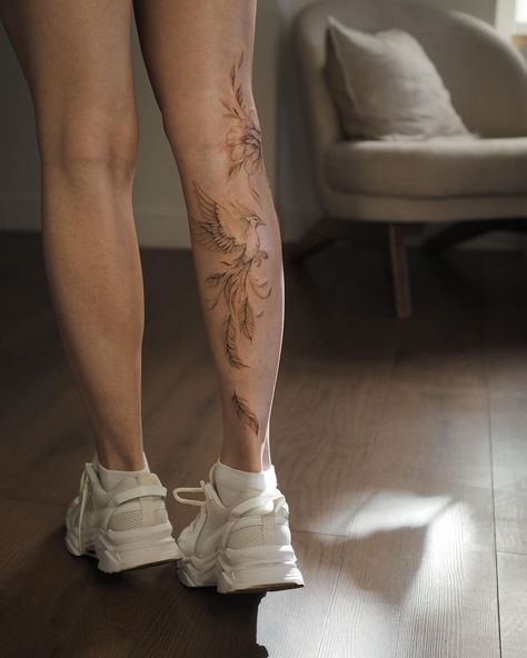 Leg with bird and floral tattoo, wearing white sneakers, standing indoors on wooden floor, near a chair.