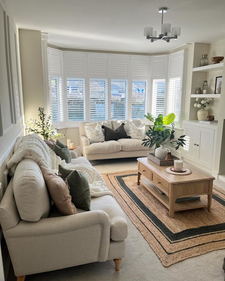 Cozy modern living room with beige sofas, wooden coffee table, plants, and large shuttered windows.