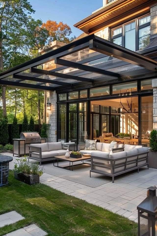 Modern patio with glass pergola, chic outdoor furniture, and lush greenery for a stylish entertaining space.