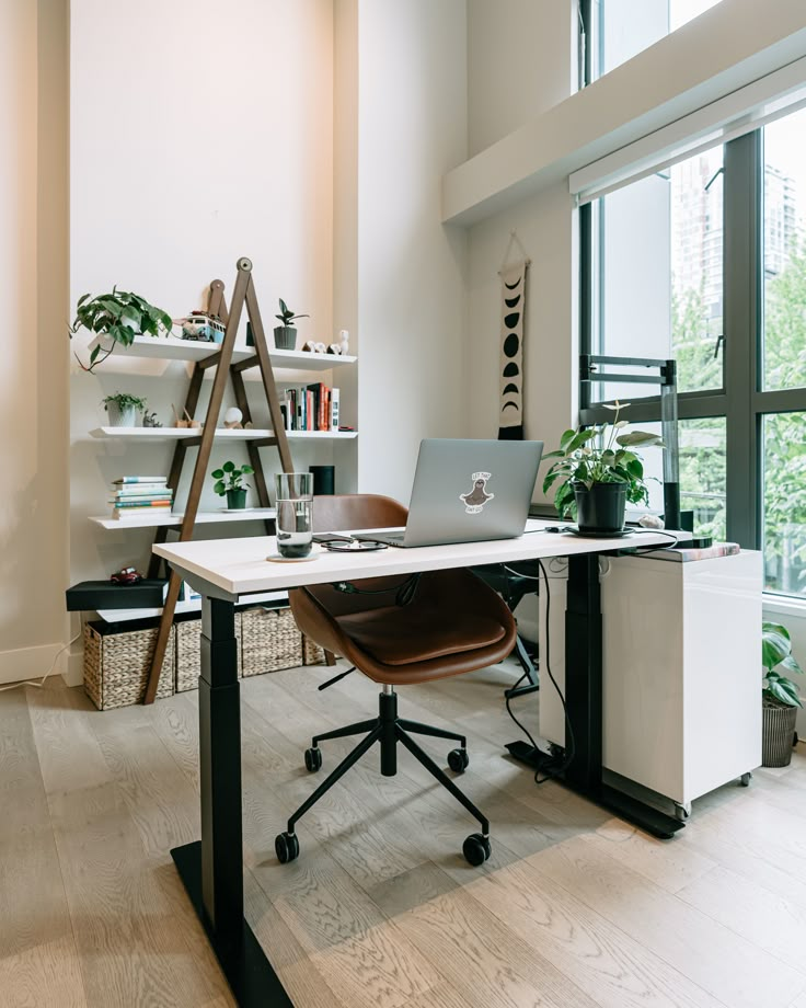 Modern home office with desk, laptop, chair, shelves, and plants by large window. Bright, organized workspace.