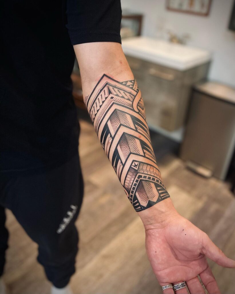 Forearm with a tribal geometric tattoo in black and gray, displayed in an indoor setting.