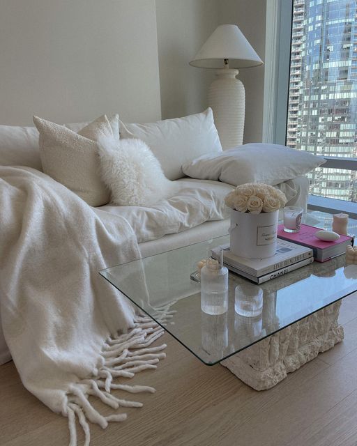 Modern cozy living room with white sofa, fluffy pillows, glass coffee table, and city view through windows.