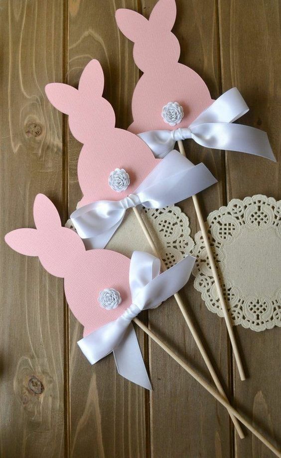 Pink bunny cutouts with white bows on sticks, placed on wooden table with lace doily. Perfect for Easter decor.