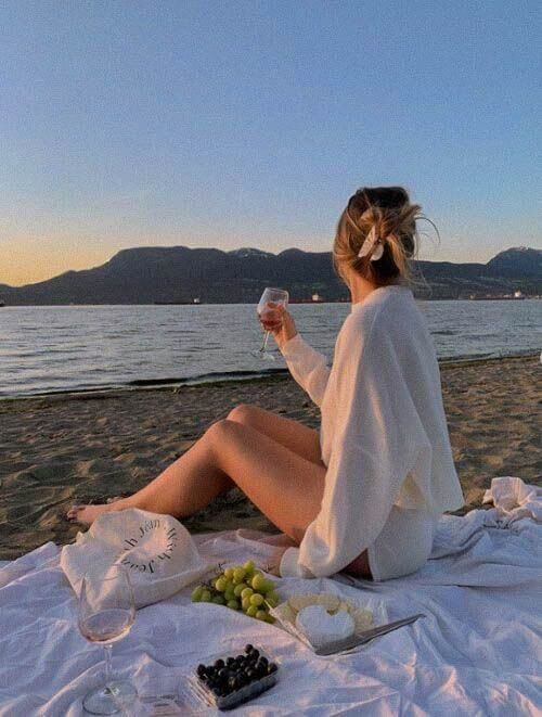 Woman enjoying a beach picnic with wine and snacks at sunset, mountains in the background.