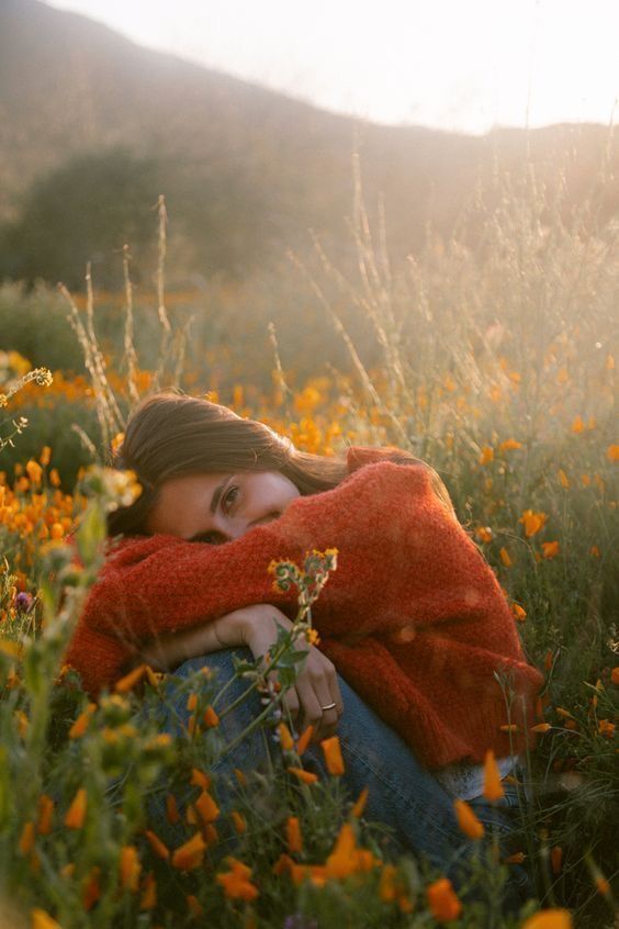 Woman in an orange sweater relaxing in a sunlit field of wildflowers at sunset, evoking tranquility and nature.